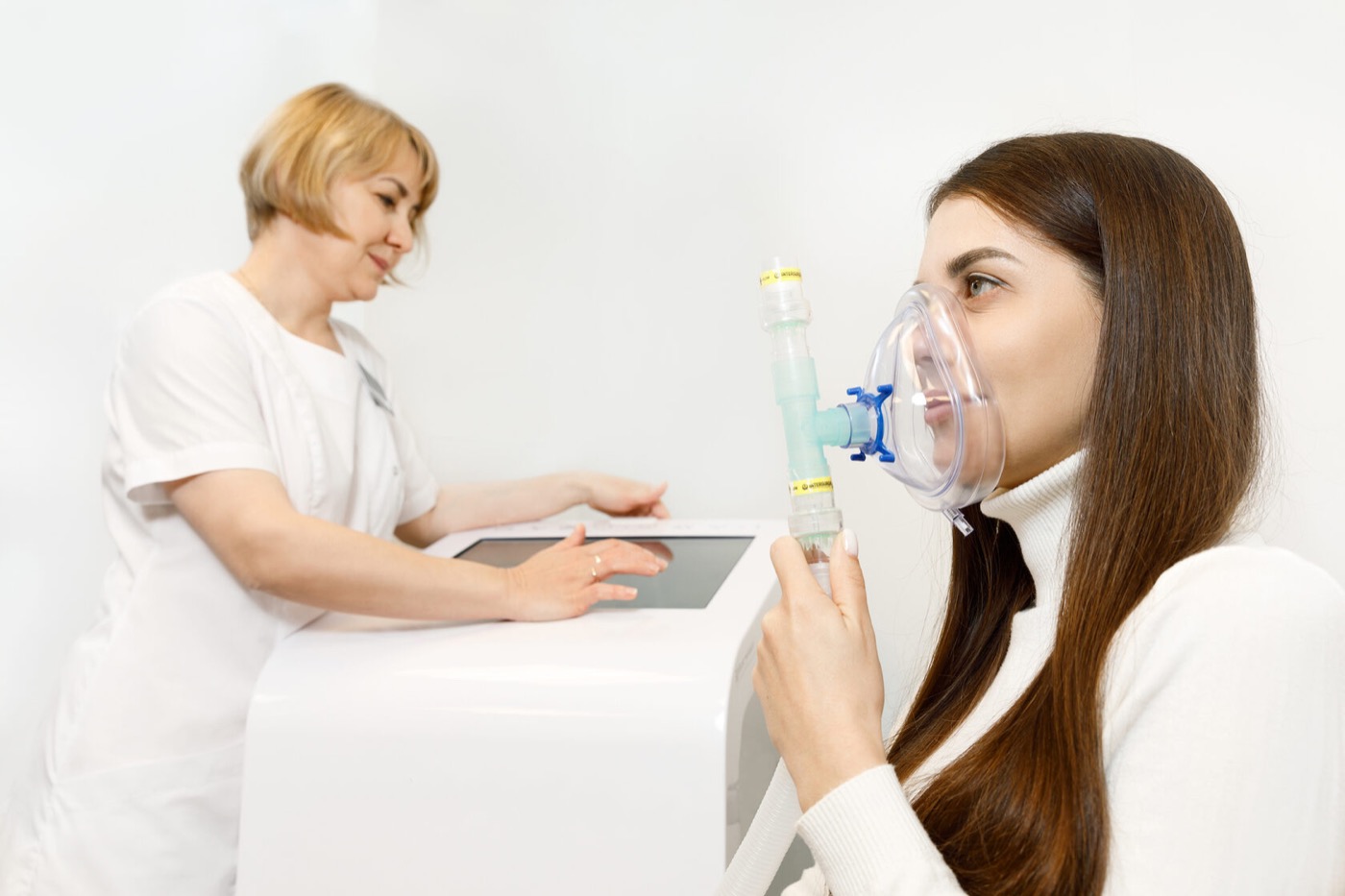 A clinician in white stands at a touchscreen console adjusting a session while a seated patient holds a clear mask to her face, connected to the console by a ribbed hose.